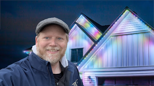 Chris Wilson stands in front of a house lit up with Gemstone lights in a soft multi coloured (light blue, light pink, yellow) alternating pattern.