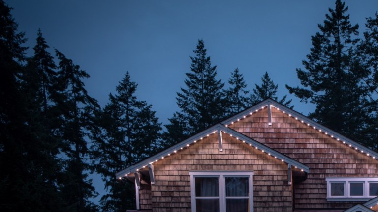 A cottage is lit up with Gemstone Lights. There are tall pine trees in the background.