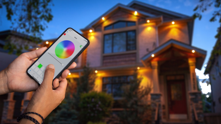 A person holds a phone with the Gemstone Lights Hub App in front of a house lit up with Gemstone lights.