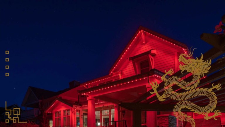 A two store house with horizontal siding has red Gemstone lights alit on all the roof lines. A gold Chinese dragon imprint is on the lower right.