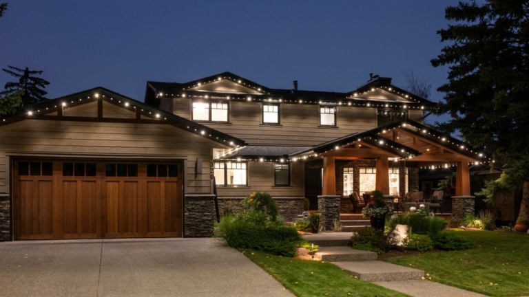A 2 storey home and garage with multiple roof lines with a combination of Gemstone regular and flood light style white lights on. Around the yard are multiple flood lights illuminated throughout the greenery. Above the picture are the words, On the Blog, the difference between uplighting and downlighting with flood lights.