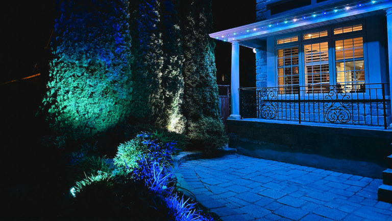 The yard of a house is illuminated with Gemstone Lights programmable outdoor flood lights in white and blue and outdoor LED track lighting in multicolour.